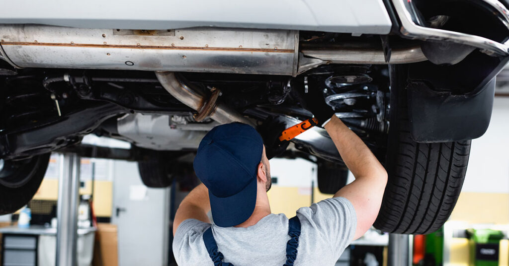 Mechanic standing underneath old car that is hoisted up as he works on replacing parts.