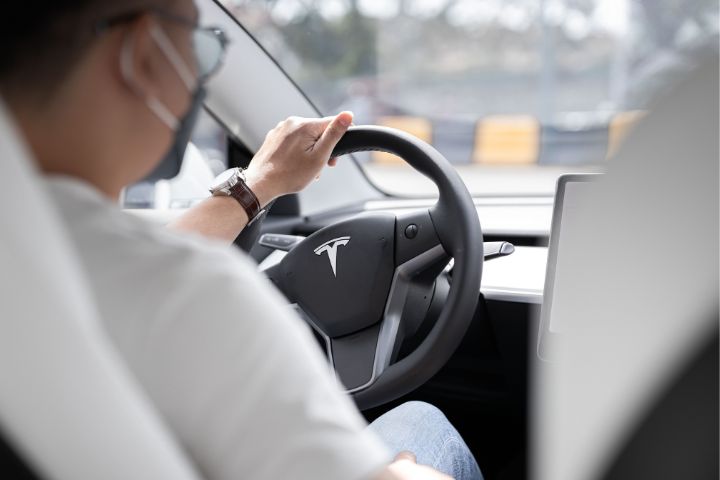 A man wearing a mask and wristwatch is driving a Tesla, with a close-up view of the minimalist interior and steering wheel bearing the Tesla logo.