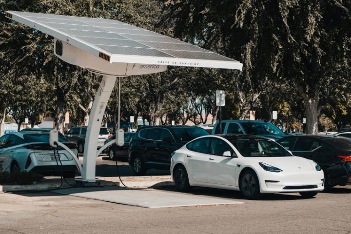A white Tesla electric car is parked at a solar-powered charging station in a parking lot surrounded by other vehicles and trees.