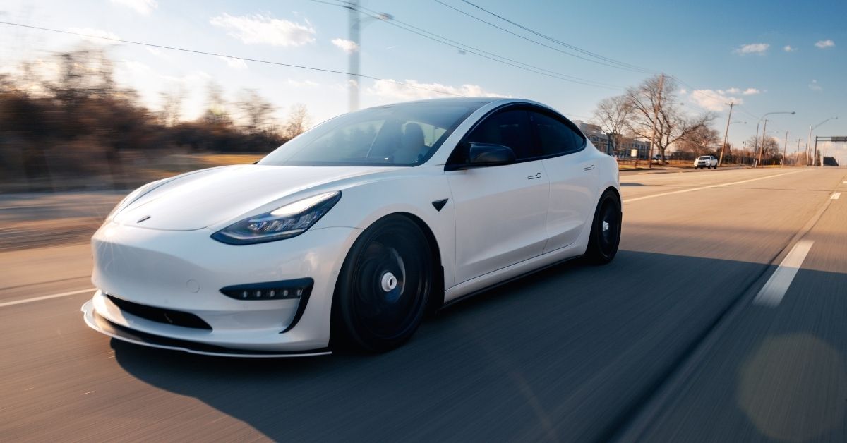 A sleek white Tesla Model 3 is driving down a sunlit, empty road with motion blur in the background.
