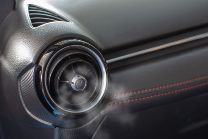 A circular car air conditioning vent is releasing cool air, with visible mist or condensation coming from the vent.