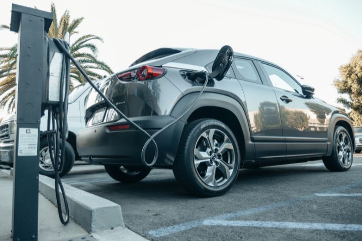 A gray electric car is parked in a charging bay, plugged into a public charging station on a sunny day.