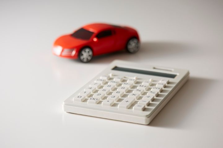 A white calculator sits on a white surface with a red toy car in the background, symbolizing car finance or budgeting.
