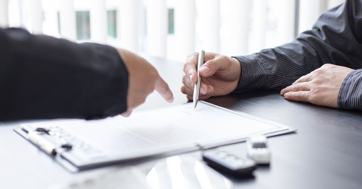 Two people sit at a table as one person points to a document while the other prepares to sign it, likely completing a used car sale, with car keys visible nearby.