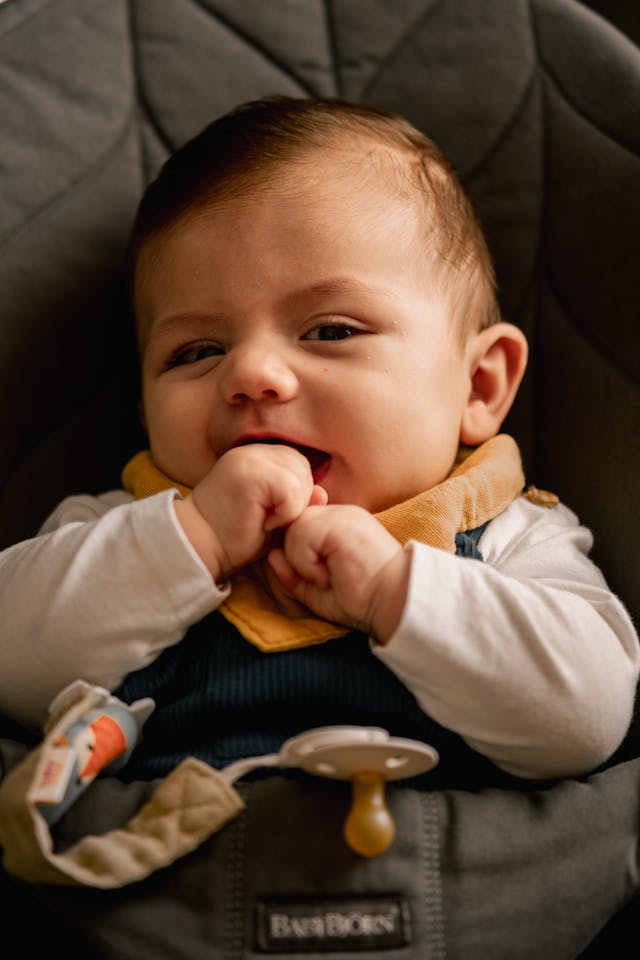 A baby sits in a car seat, smiling and chewing on their fingers, with a pacifier clipped to their outfit.