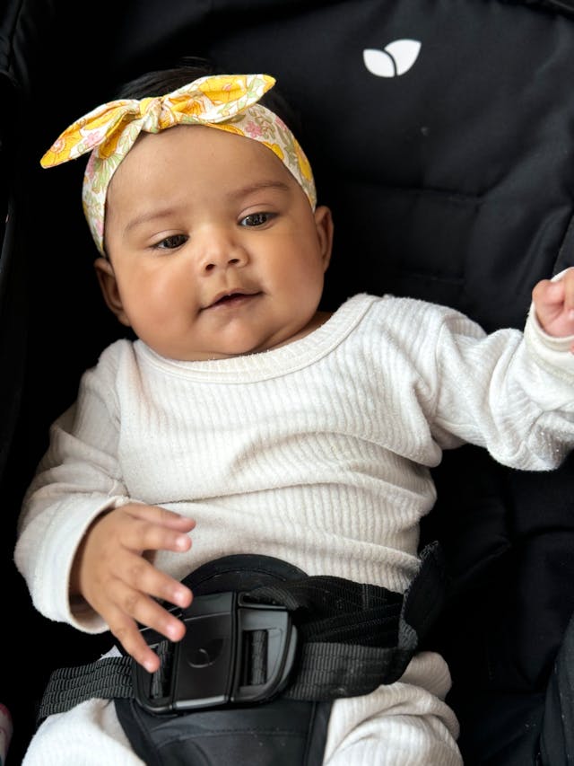 A baby wearing a white outfit and a floral headband sits secured in a car seat with a black seatbelt.