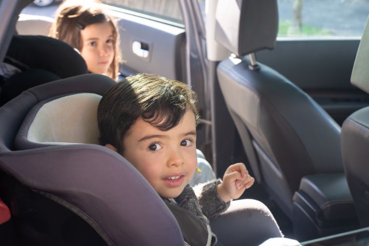 A young boy sits in a car seat looking toward the camera, with a girl seated behind him in the back seat.