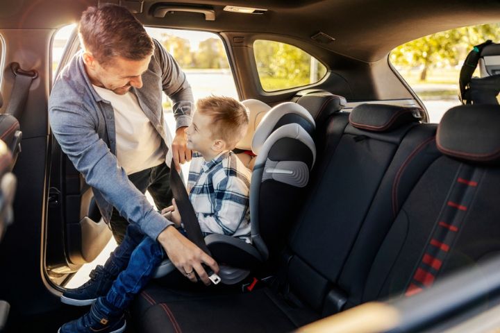 A man helps buckle a young boy into a booster seat in the back of a car, both smiling at each other.