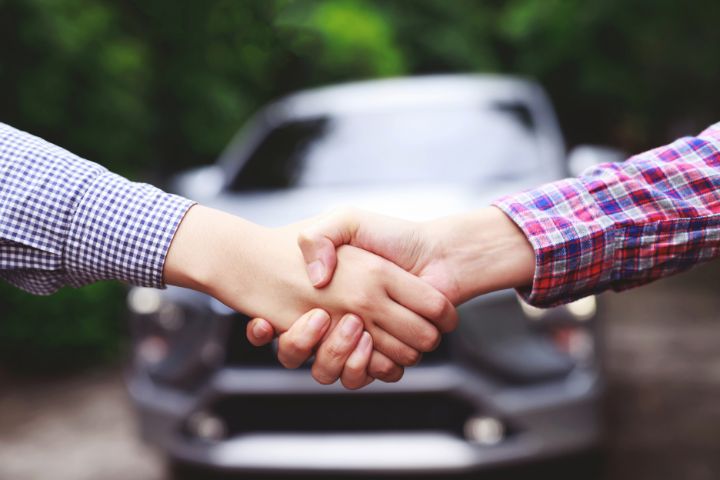 Two people shake hands in front of a car, symbolizing the completion of a deal, likely the buying or selling of a used vehicle.