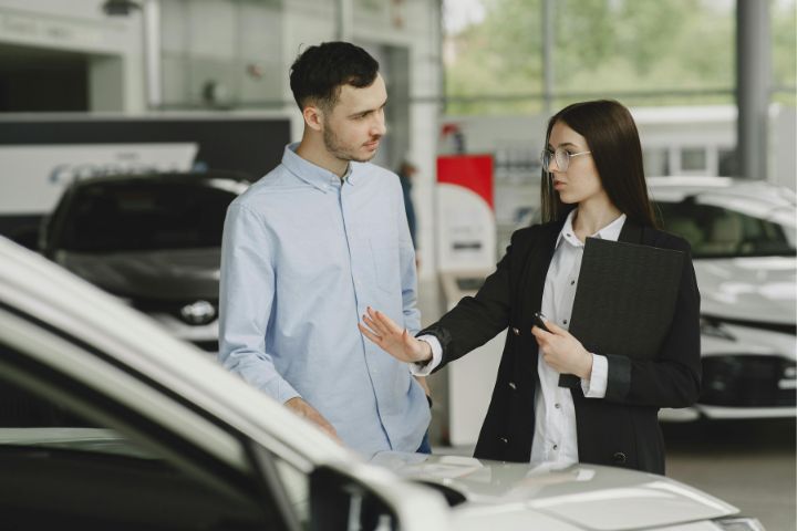 A car saleswoman is explaining details about a vehicle to a potential customer inside a showroom, gesturing toward the car while holding a folder.